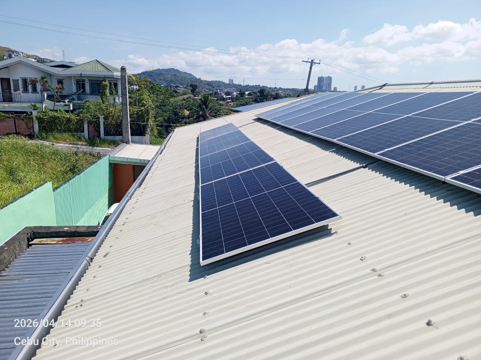 Panels arranged on a white corrugated rooftop in a Cebu City hillside neighborhood, a neighbor's home anchoring the foreground and the city skyline reaching into the distance.