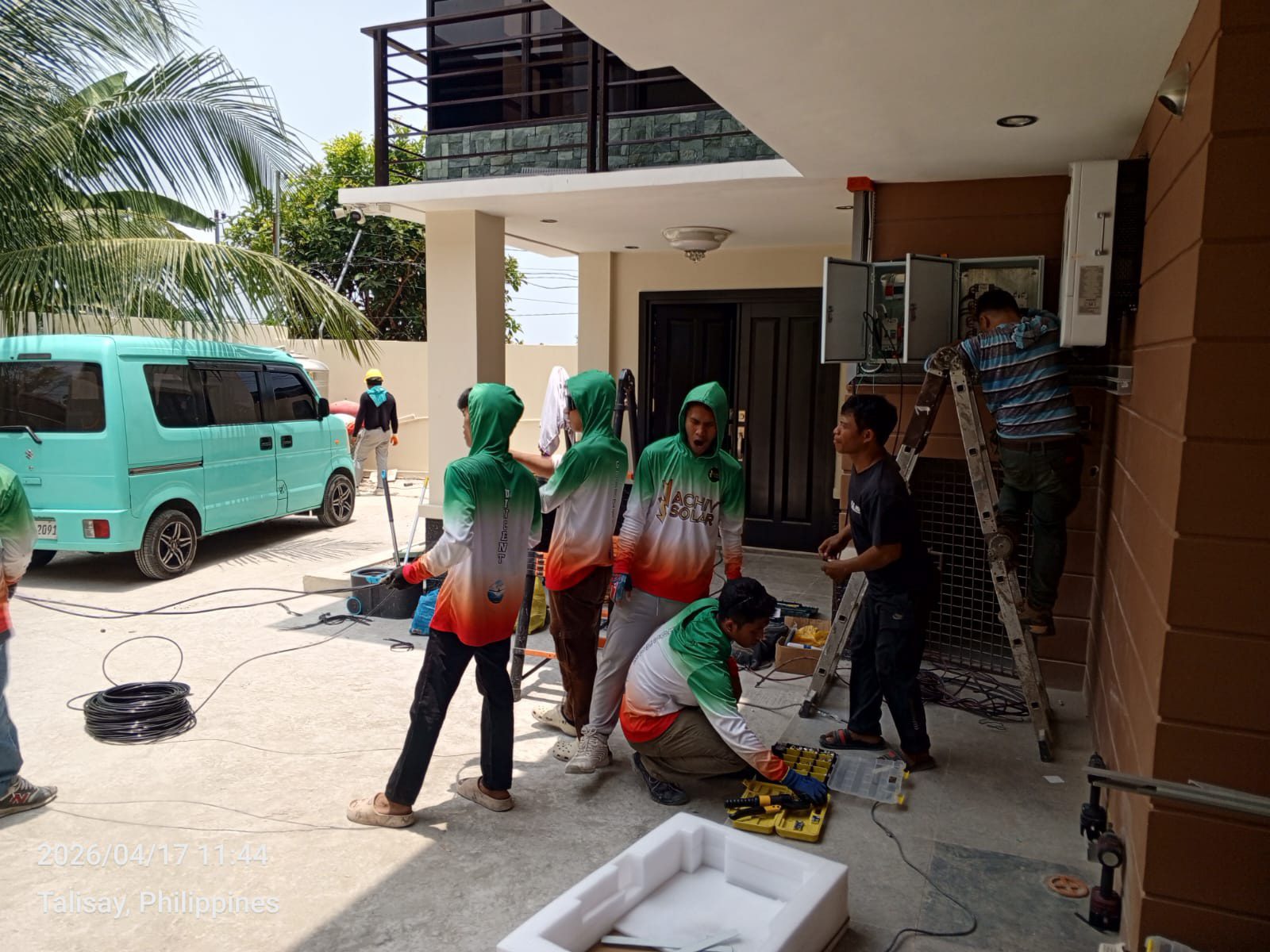 Six-person Achiv install team staging the inverter cabinet at the front door of a Talisay home on install day, teal van parked, ladder and tools spread across the driveway.
