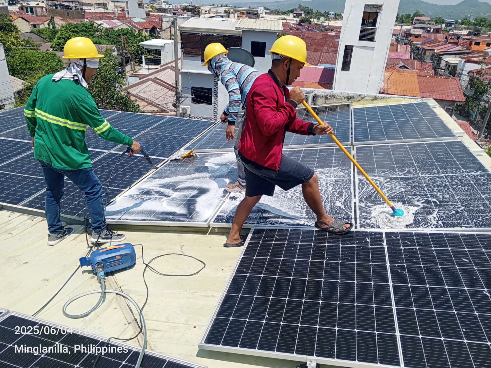 Three Achiv cleaners in hardhats and hi-vis vests applying soapy foam and mopping a residential panel array above a Minglanilla neighborhood.