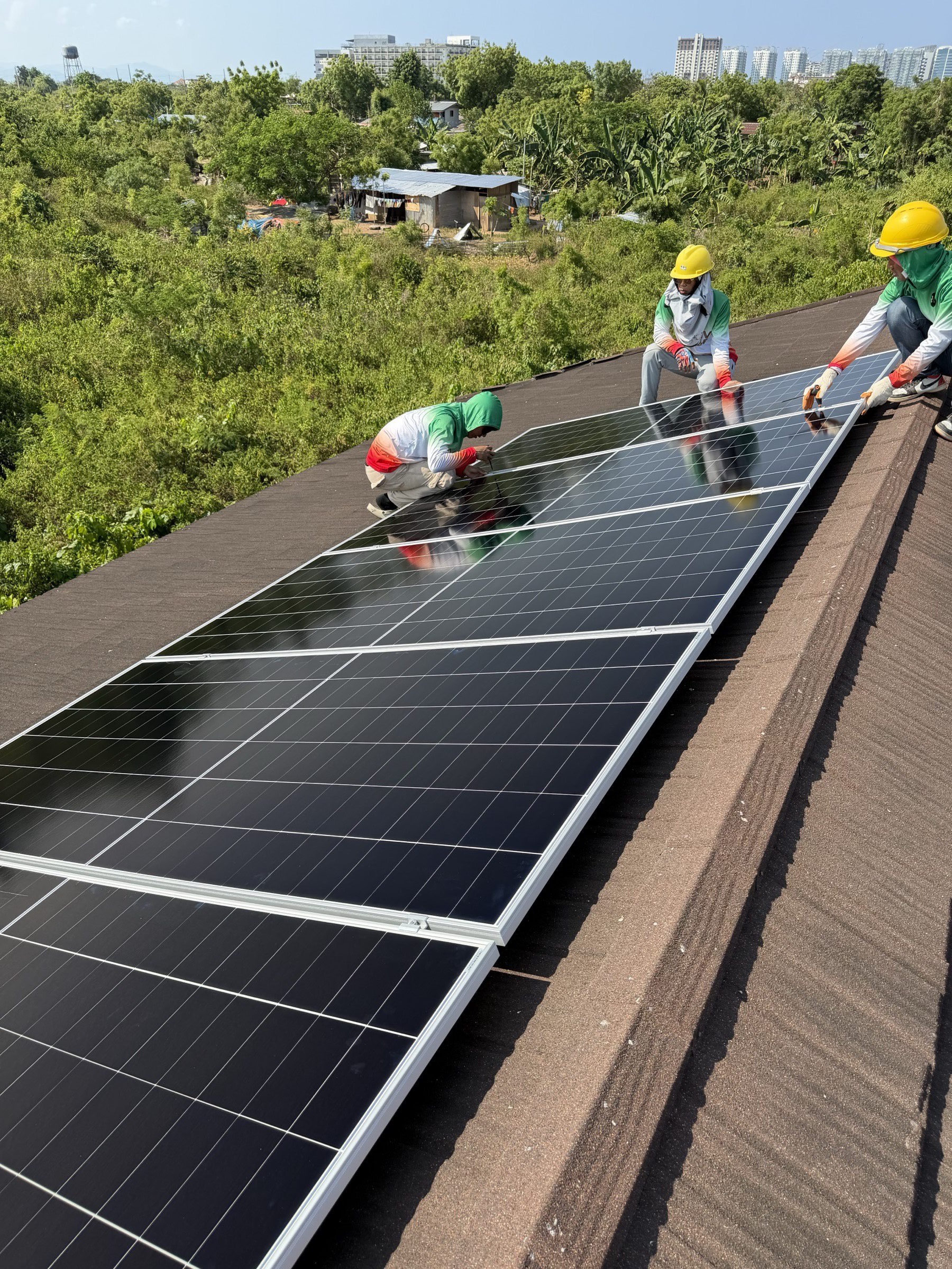 Three Achiv Solar technicians installing a long panel array on a Filipino residential rooftop, lush hillside and city skyline behind them under a deep blue sky.